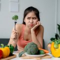 An unimpressed woman holding up a piece of broccoli on a fork.